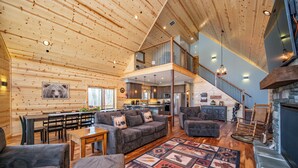 Un vaste espace de vie doté d’un plafond haut, de murs en bois, d’un coin salon central avec des canapés et une table basse, d’une cheminée et d’une cuisine à l’arrière-plan.