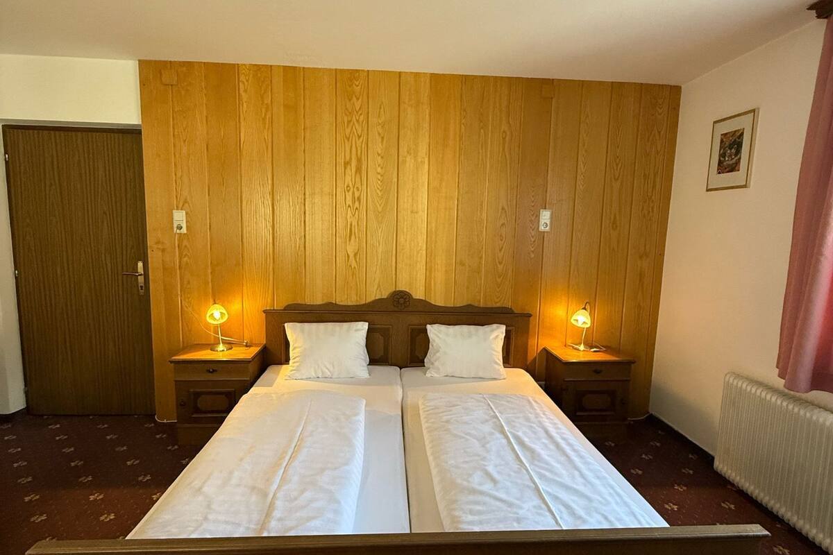 A double bed with wooden headboard, two bedside tables with lamps, a wooden panel wall, a door, and a radiator.