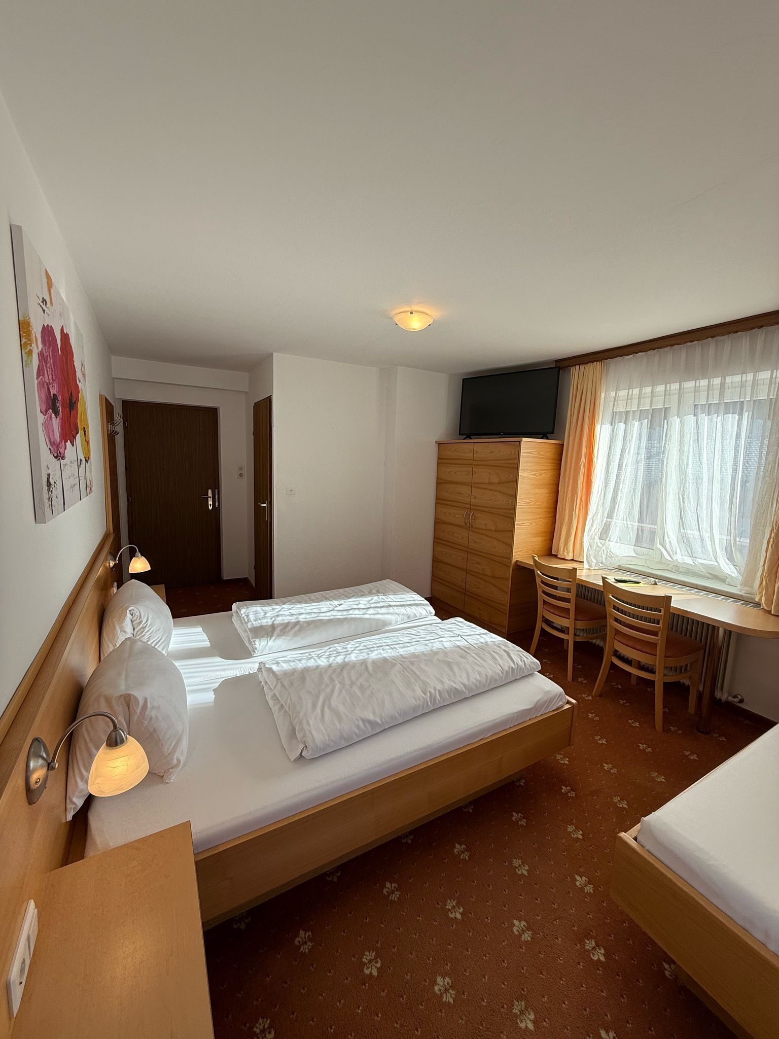 Una camera d'hotel con un letto, comodini, una scrivania e una finestra con tende leggere.