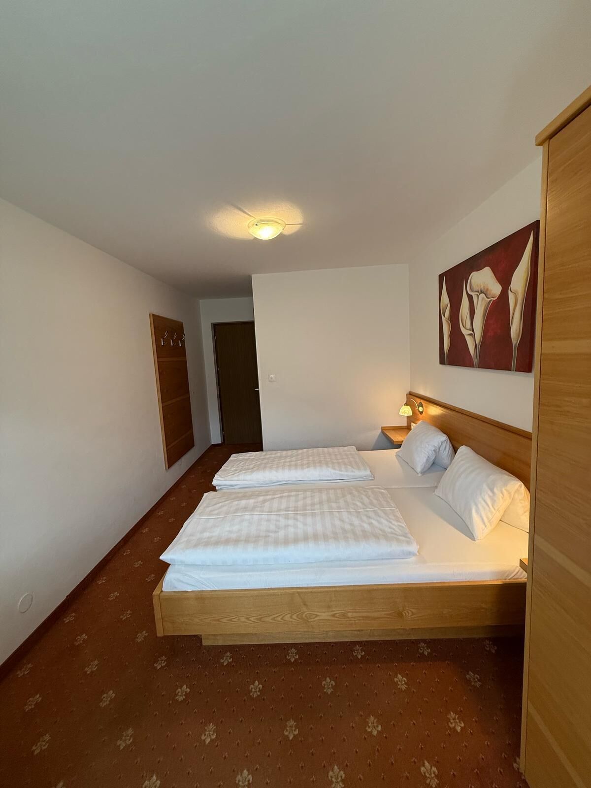 A hotel room with a large bed, a wooden headboard, a bedside table with a lamp, a door, and a wall-mounted artwork.