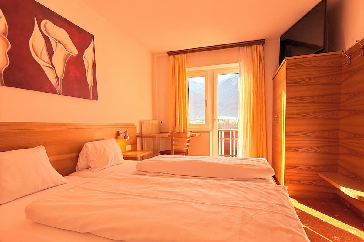 A hotel room with a large bed, a wooden headboard, a painting on the wall, a window with curtains, and a balcony.