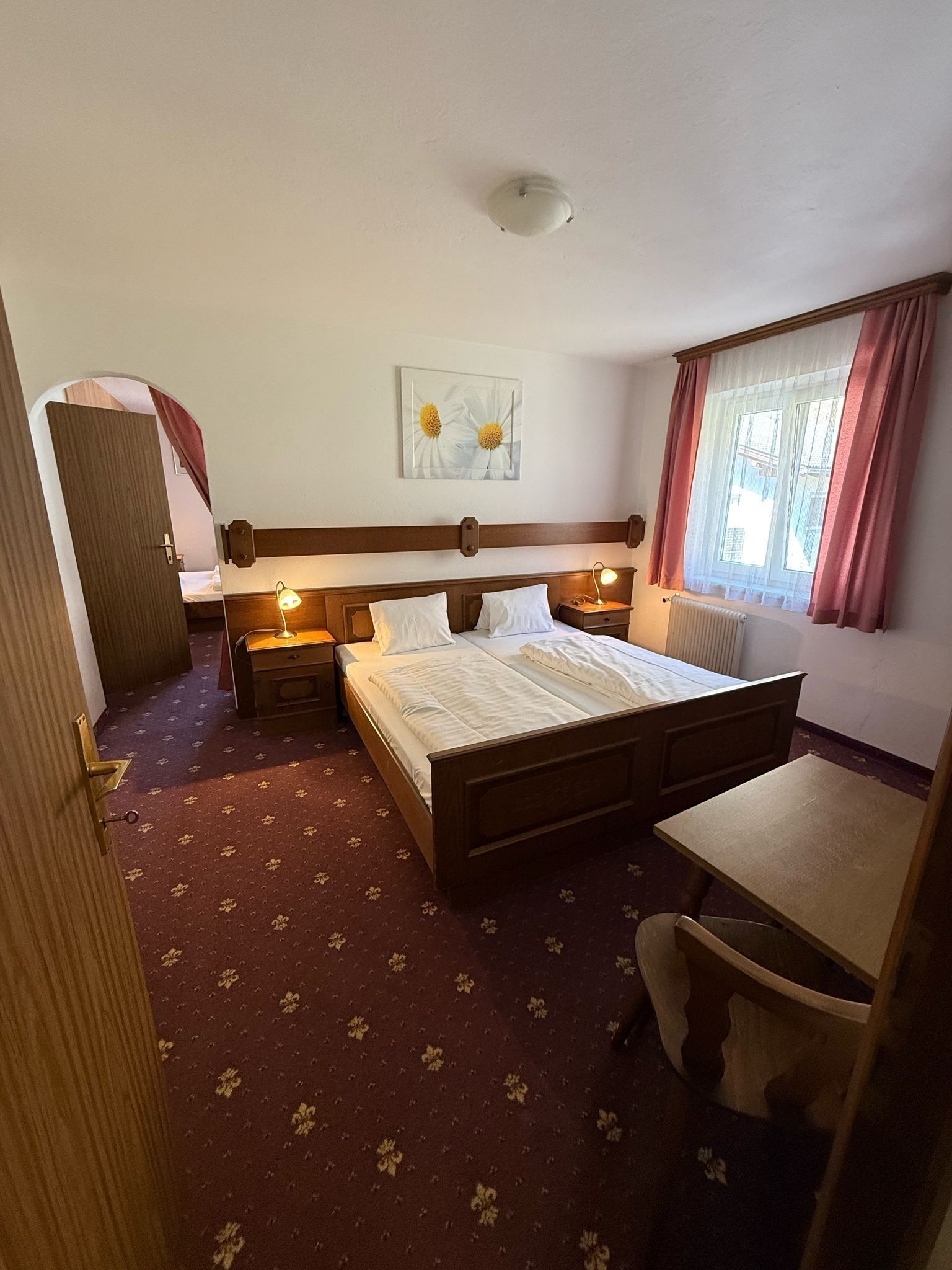 Ein Hotelzimmer mit einem Bett, Nachttischen, einem Schreibtisch, einem Sessel, einer Heizung, einem Fenster mit Vorhängen und einer Tür.