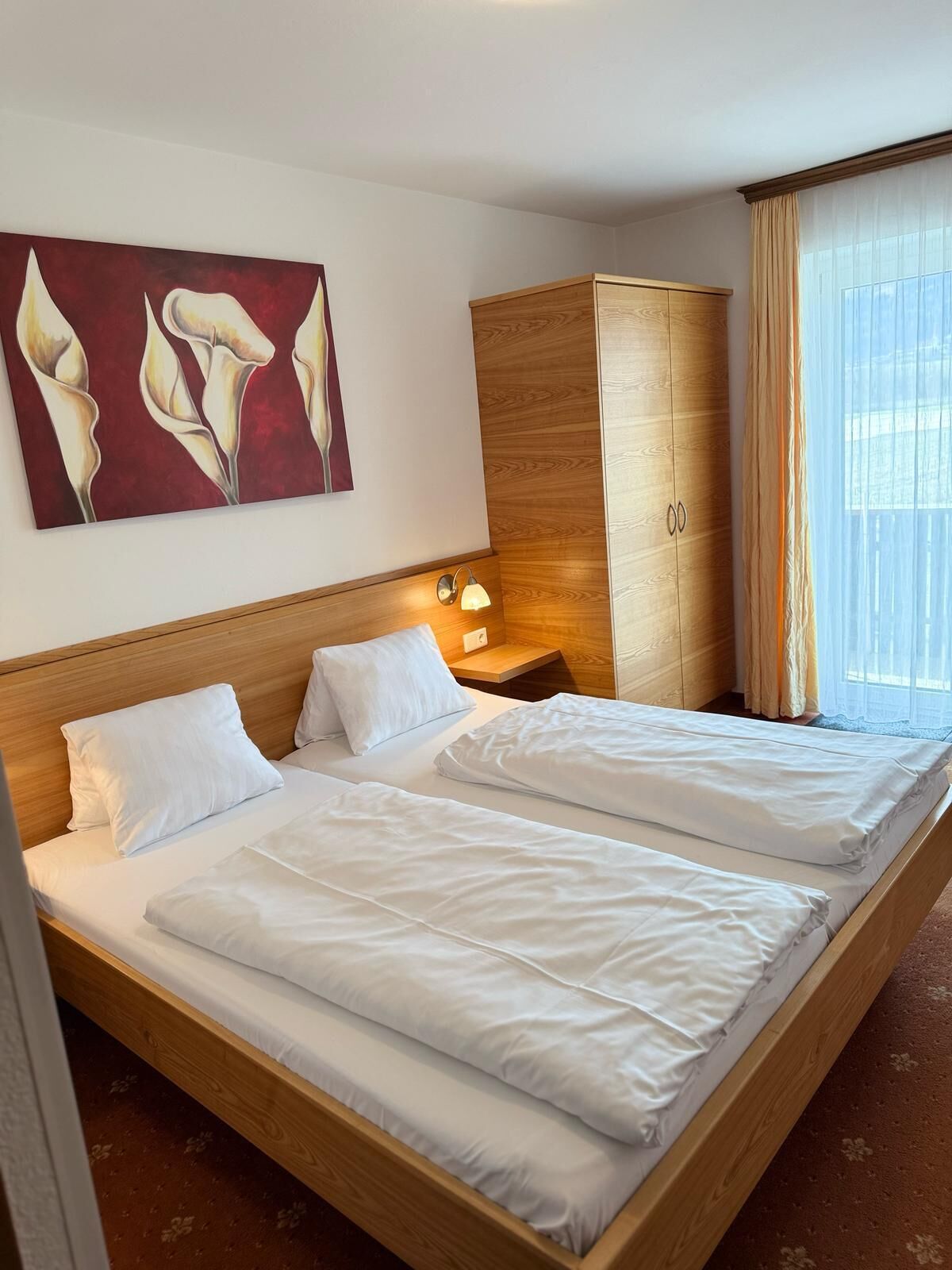 A hotel room with two beds, a wooden wardrobe, a nightstand with a lamp, and a painting on the wall.
