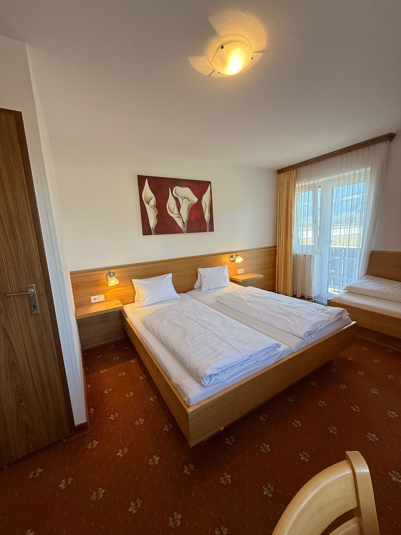 A hotel room with a large bed, two bedside tables with lamps, a ceiling light, a window with curtains, and a floral carpet.
