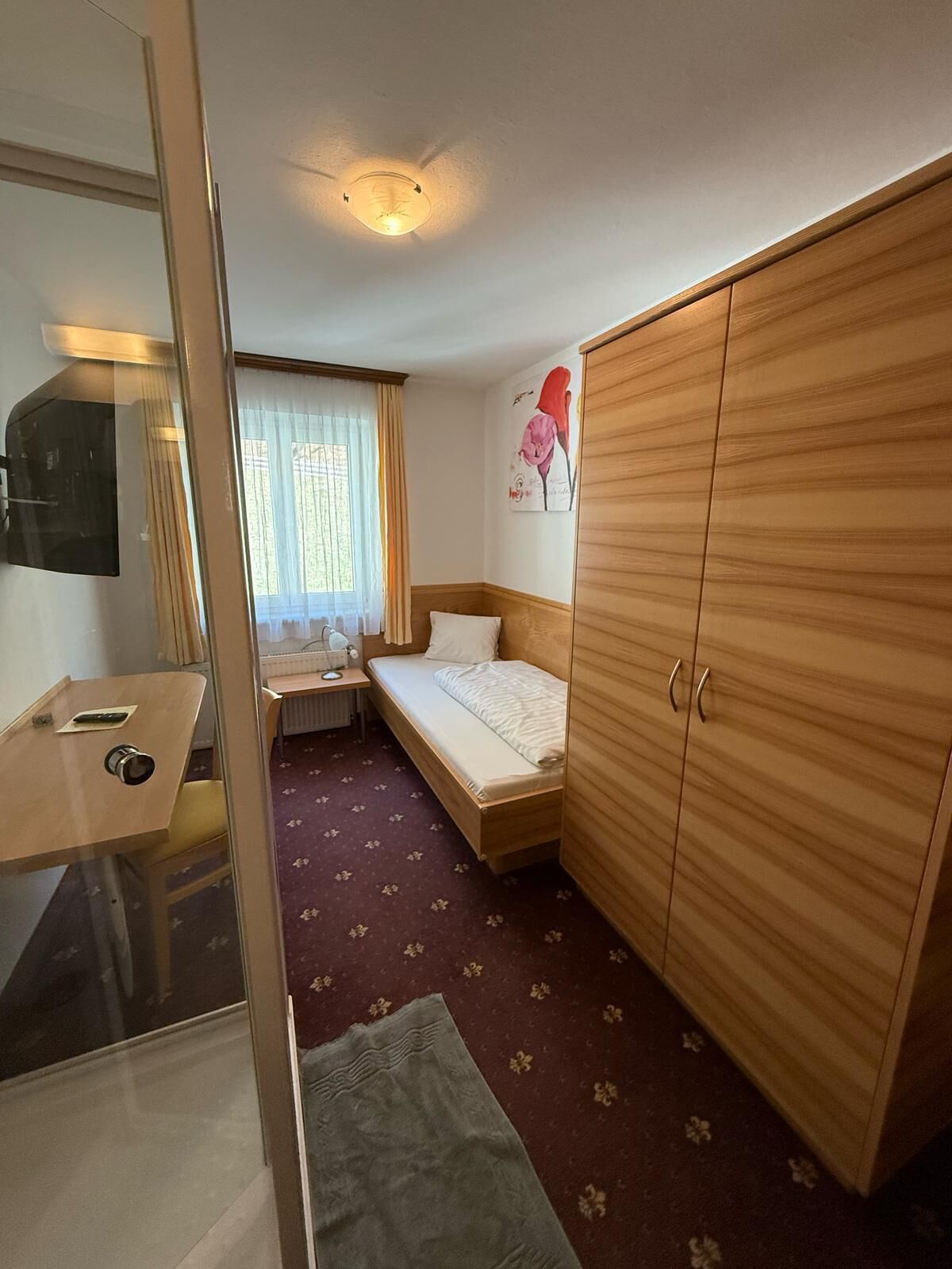 A compact hotel room with a single bed, a wooden wardrobe, a desk with a chair, a small table, and a window with curtains.