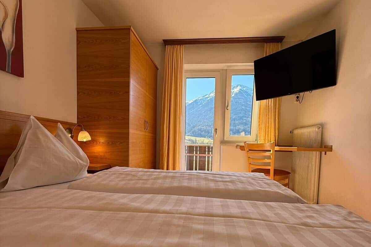 A hotel room with a large bed, a television mounted on the wall, a wooden wardrobe, and a view of mountains through the window.