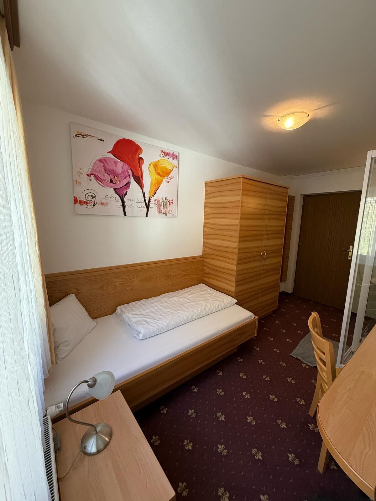 A compact room with a bed, a wooden wardrobe, a desk with a lamp, and a dining table.