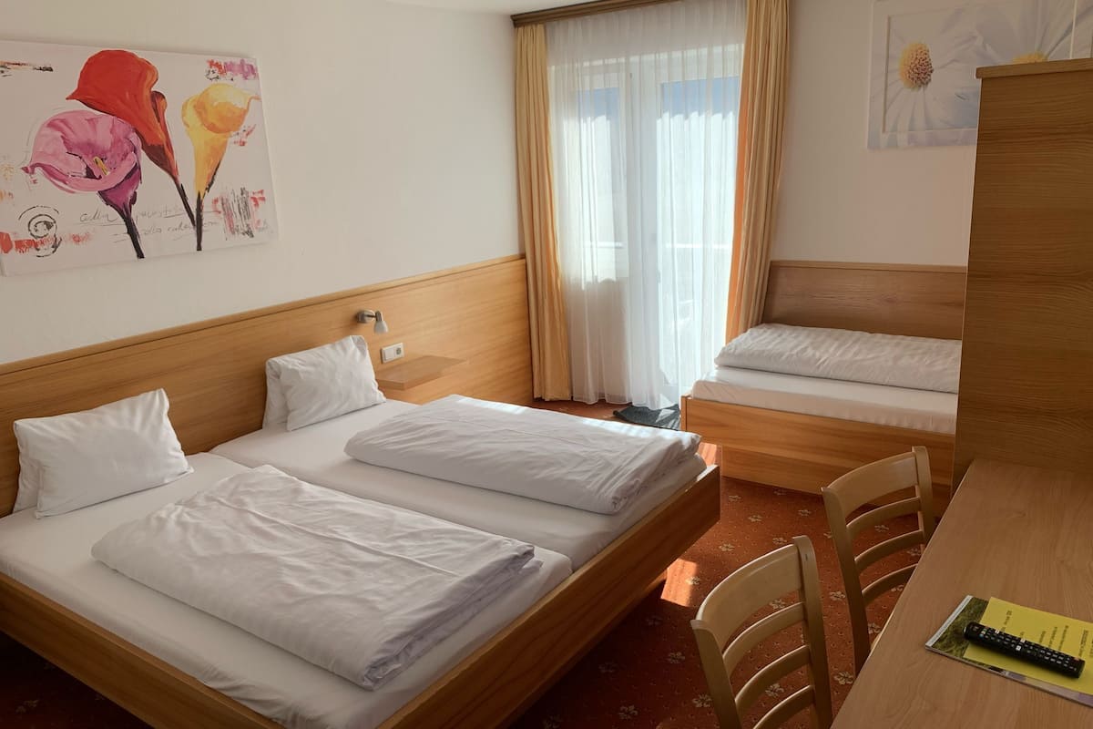 A hotel room with two beds, a wooden headboard, a nightstand, a lamp, a window with sheer curtains, a painting of flowers, and a wooden desk with a remote control.