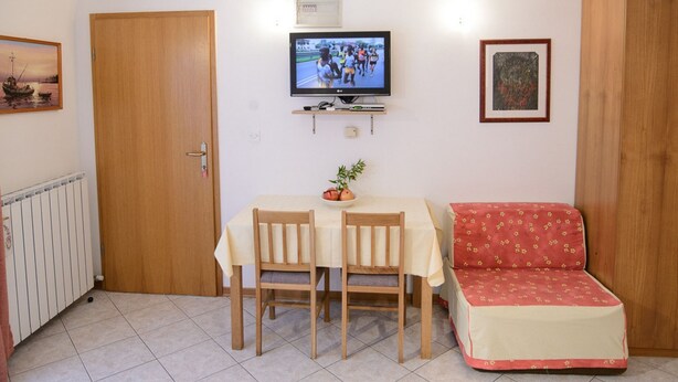 Ένα μικρό σαλόνι με μια τηλεόραση, ένα τραπέζι τραπεζαρίας με καρέκλες, έναν καναπέ και έναν πίνακα σε κορνίζα στον τοίχο.