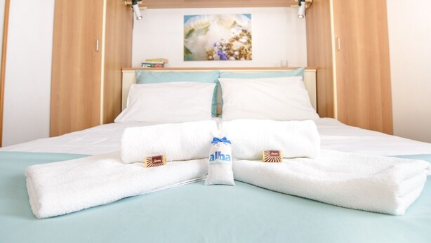 A neatly made bed with white linens and a folded towel, a small table with books, and a painting on the wall. - Sunny Street Apartments (Rovinj)