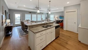 A modern kitchen with a granite countertop, white cabinetry, and a central island. There are two pendant lights, a ceiling fan, and a dining area with a table and chairs. - Abigail's House, 3 bedroom 2 bath home sleeps10. A short stroll to the beach. (Crystal Beach)