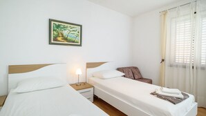 Une chambre d’hôtel avec deux lits, une table de chevet avec une lampe, une chaise, une fenêtre avec des rideaux et un tableau au mur.