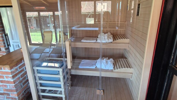 Eine Sauna mit Holzbänken und Glastüren, einer Uhr sowie einem sichtbaren Außenbereich mit Ziegelwand und einer Leiter.