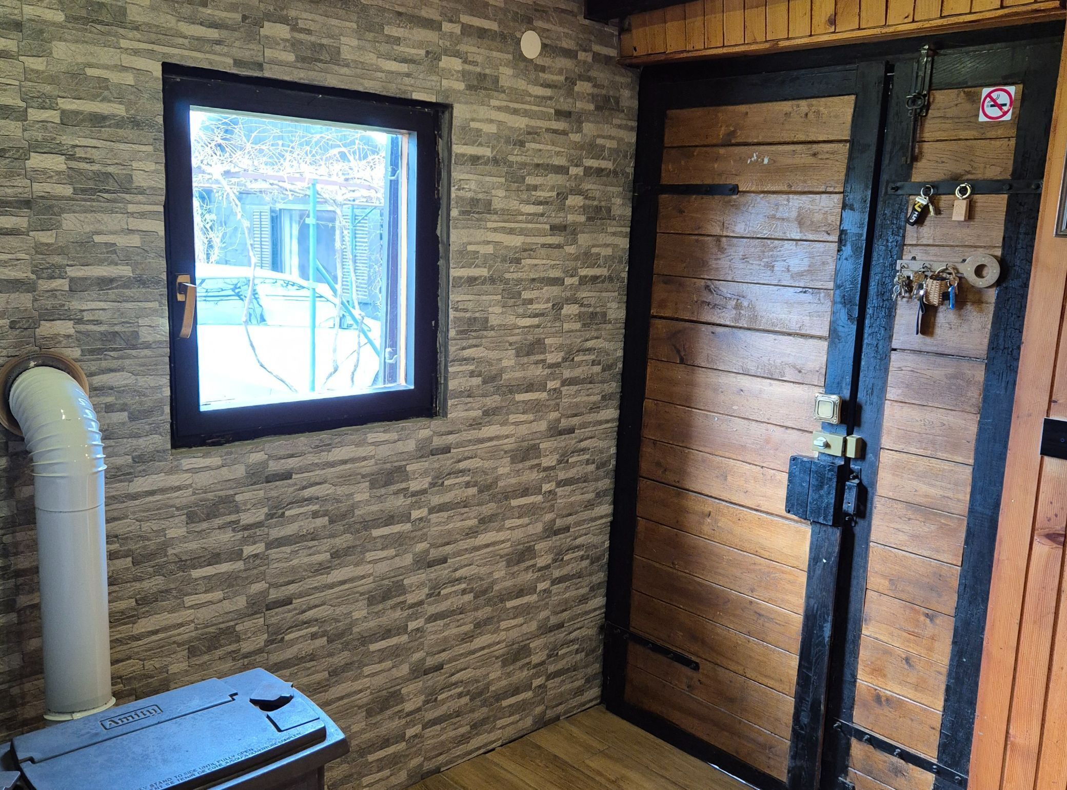 A room with a stone wall, a window with a view, a wooden door with a lock, and a small table with a chair.