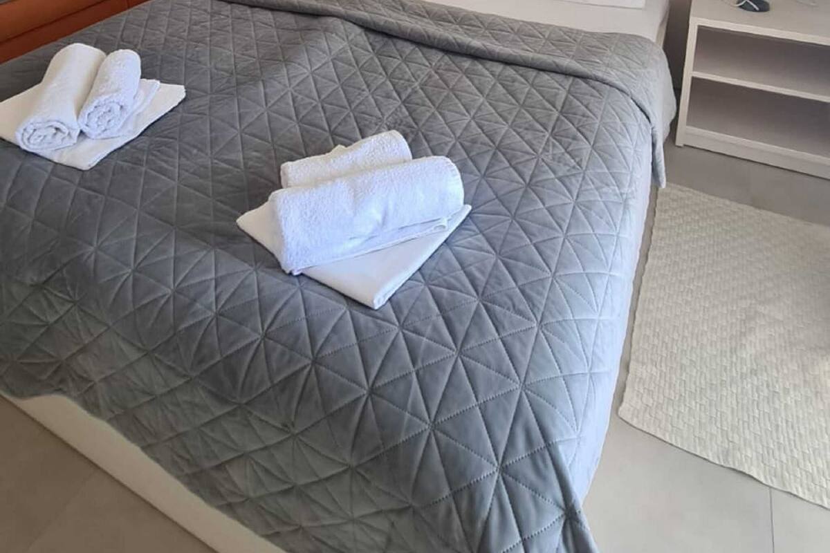 A neatly made bed with a grey quilt, two white pillows, and folded white towels. There is a white nightstand with a lamp, a small table with a lamp, and a large mirror on the wall.