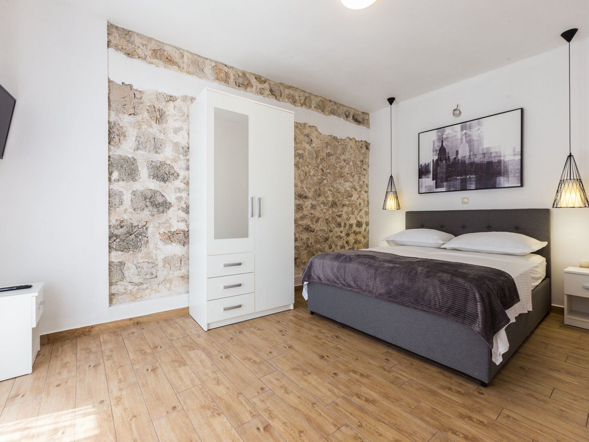 A bedroom with a stone wall, a bed with a gray headboard, a white wardrobe, and a framed picture on the wall.