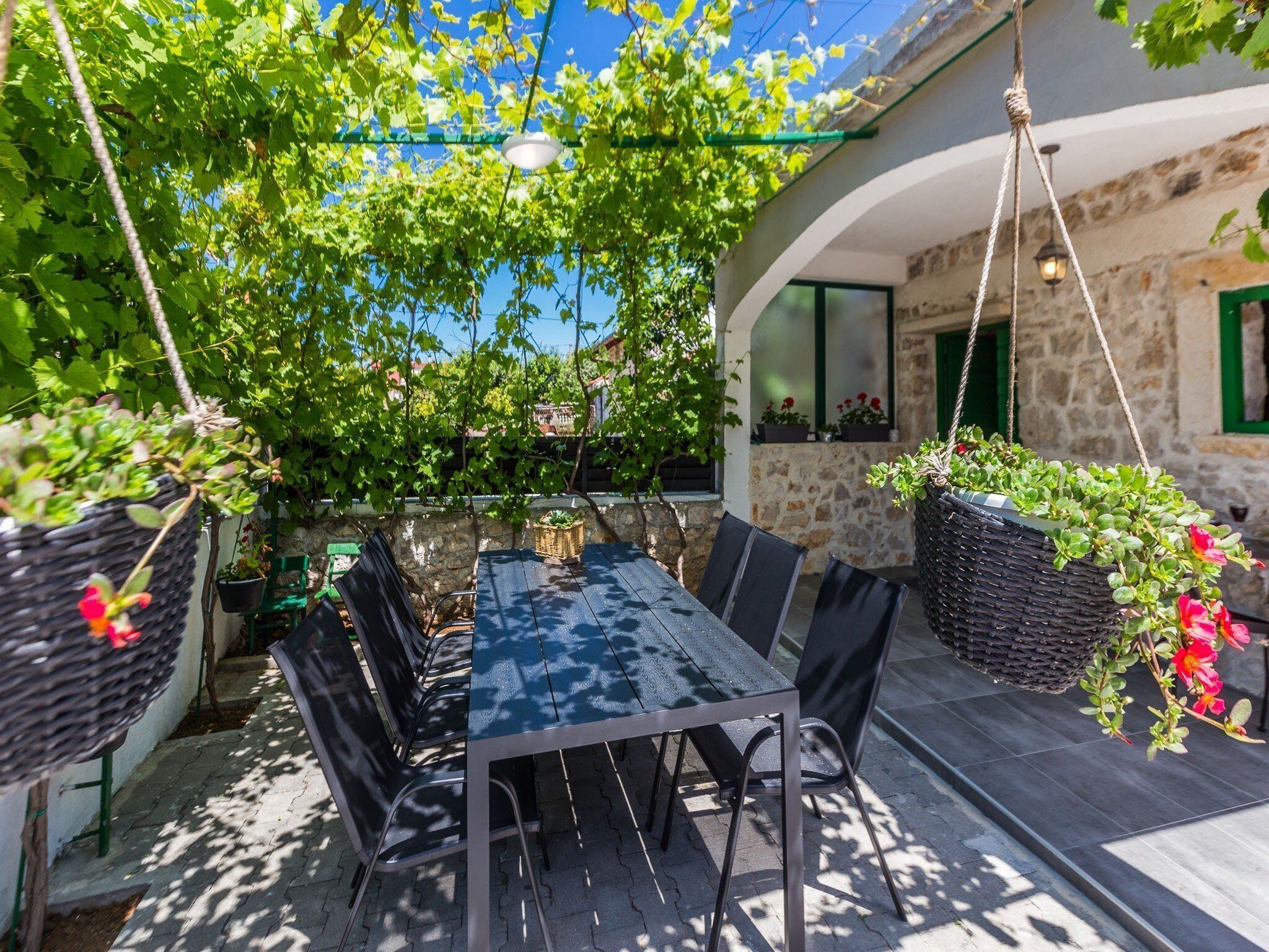 An outdoor dining area with a table and chairs, surrounded by greenery and a stone building.