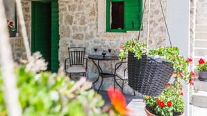 Ein stattliches Steinhaus mit rustikalem Charme, mit einer kleinen Sitzterrasse, einem Tisch und Stühlen, einer Hängetasche mit Blumen und einer grünen Tür.