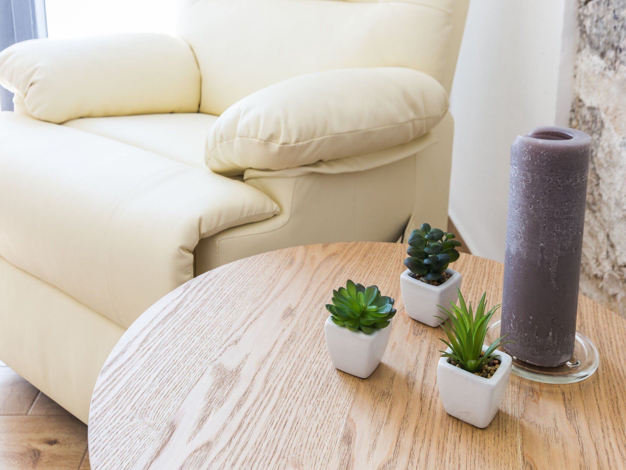 Een beige bank, een ronde houten tafel met drie kleine planten in potten, en een hoge, cilindervormige object.
