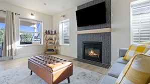 A modern living room with a fireplace, a television mounted on the wall, and a comfortable seating area with a sofa and ottoman. - Beautiful Lakefront Home with Spa and 6 Bedrooms! (Moses Lake)