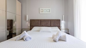 Een net opgemaakt bed met witte lakens en twee opgerolde handdoeken, een grote spiegel en een ingelijst foto aan de muur.