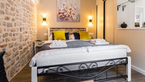 A bedroom with a stone wall, a bed with white and gray bedding, two yellow pillows, and a floral painting above the bed. - Authentic Stay in the Heart of Zadars Old Town (Zadar)