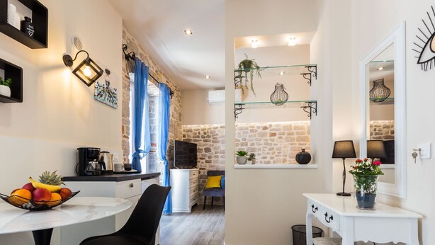 A modern kitchen with a stone wall, a dining area with a white table and black chairs, and a small white console table with a vase of flowers. - Authentic Stay in the Heart of Zadars Old Town (Zadar)