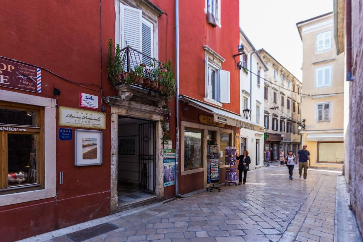 A narrow cobblestone street with a red building on the left and a yellow building on the right. There are storefronts with signs for a barber shop and a hotel.