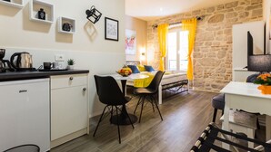 A compact kitchen with a coffee maker, kettle, and a white refrigerator. A dining area with a table and chairs is adjacent to a bed with yellow curtains. - Authentic Stay in the Heart of Zadars Old Town (Zadar)