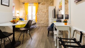 A cozy room with a stone wall, a dining table with chairs, a TV, and a desk with a lamp. - Authentic Stay in the Heart of Zadars Old Town (Zadar)