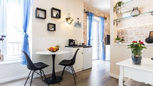 A modern kitchen with a white countertop, a coffee machine, and a small refrigerator. There is a round table with two chairs and a bowl of fruit on it. The room has a stone wall and blue curtains. - Authentic Stay in the Heart of Zadars Old Town (Zadar)