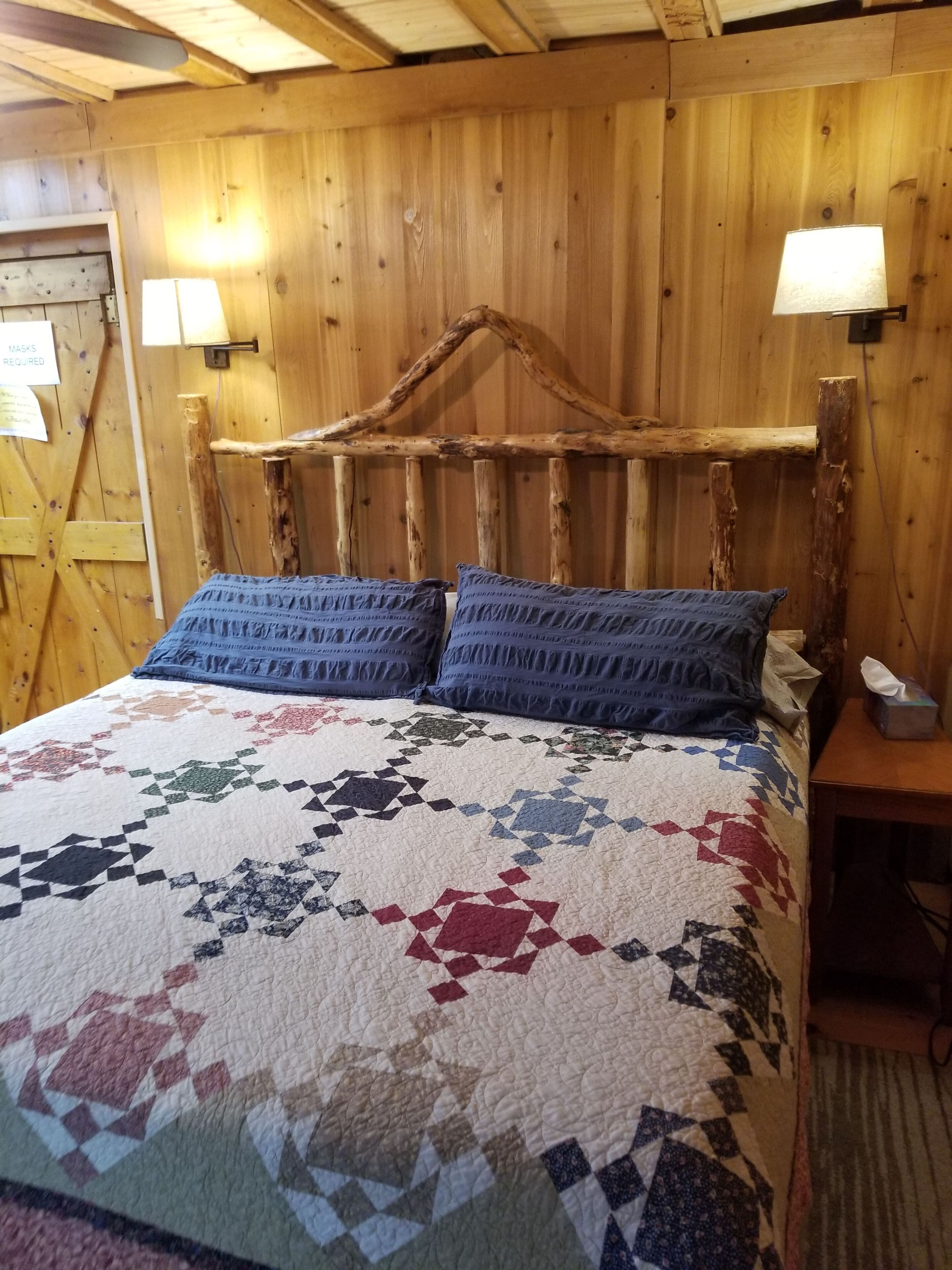 A wooden cabin room with a bed, two wall-mounted lamps, and a sign indicating mask requirements.