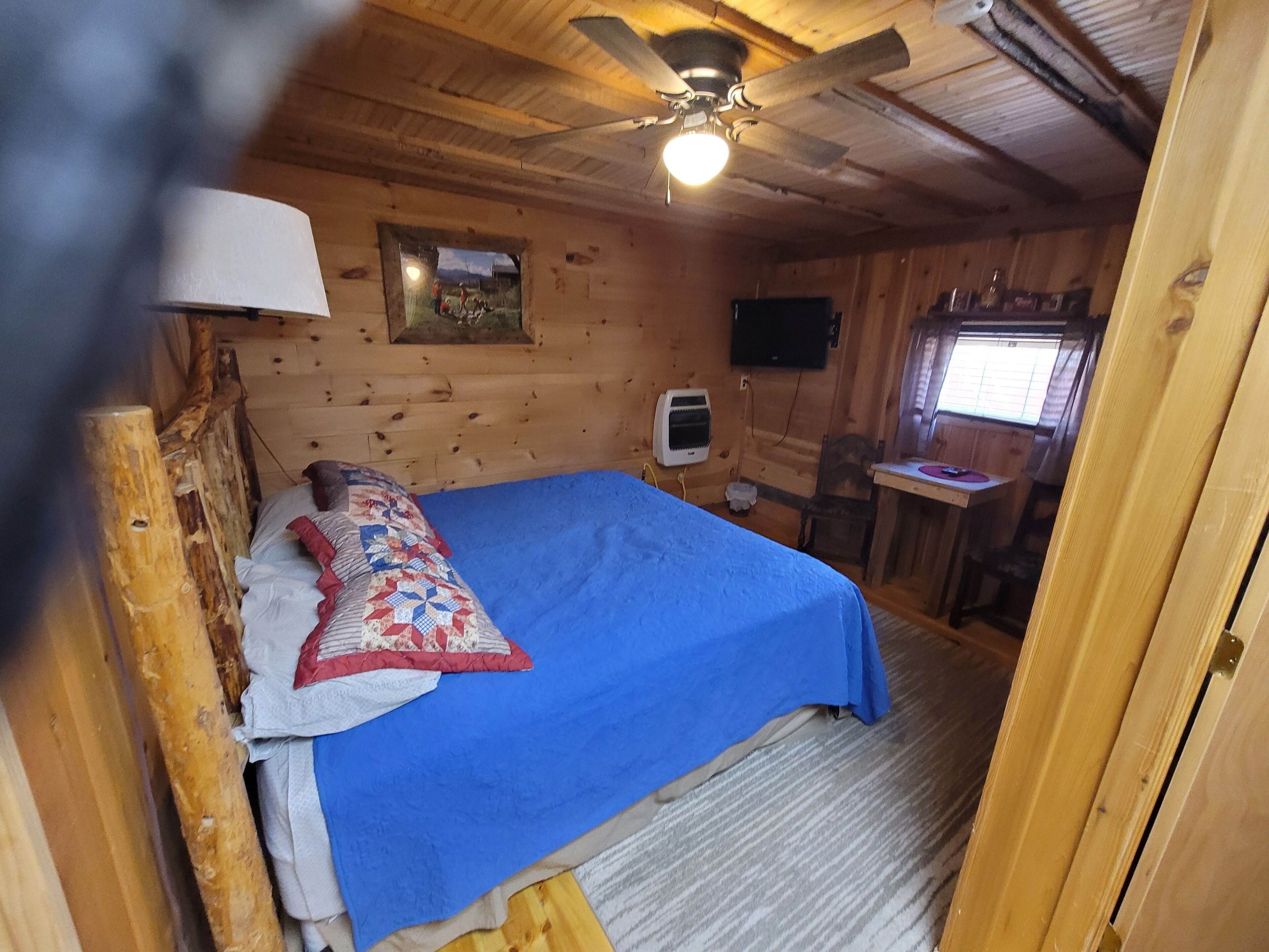 Ein Zimmer in einer Holzhütte mit einem Bett, einem Deckenventilator, einem kleinen Schreibtisch und einem Fenster.