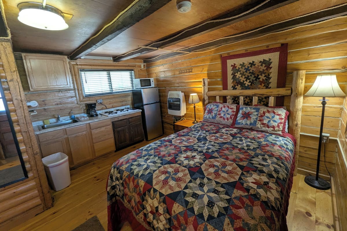 A cozy cabin interior with a wooden interior, a bed with a quilt, a kitchenette, and a window with blinds.