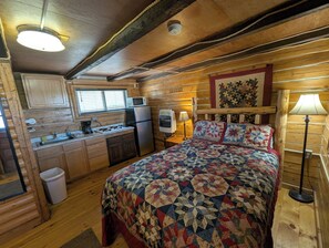 A cozy cabin interior with a wooden interior, a bed with a quilt, a kitchenette, and a window with blinds. - CopperLine Lodge (Saratoga)