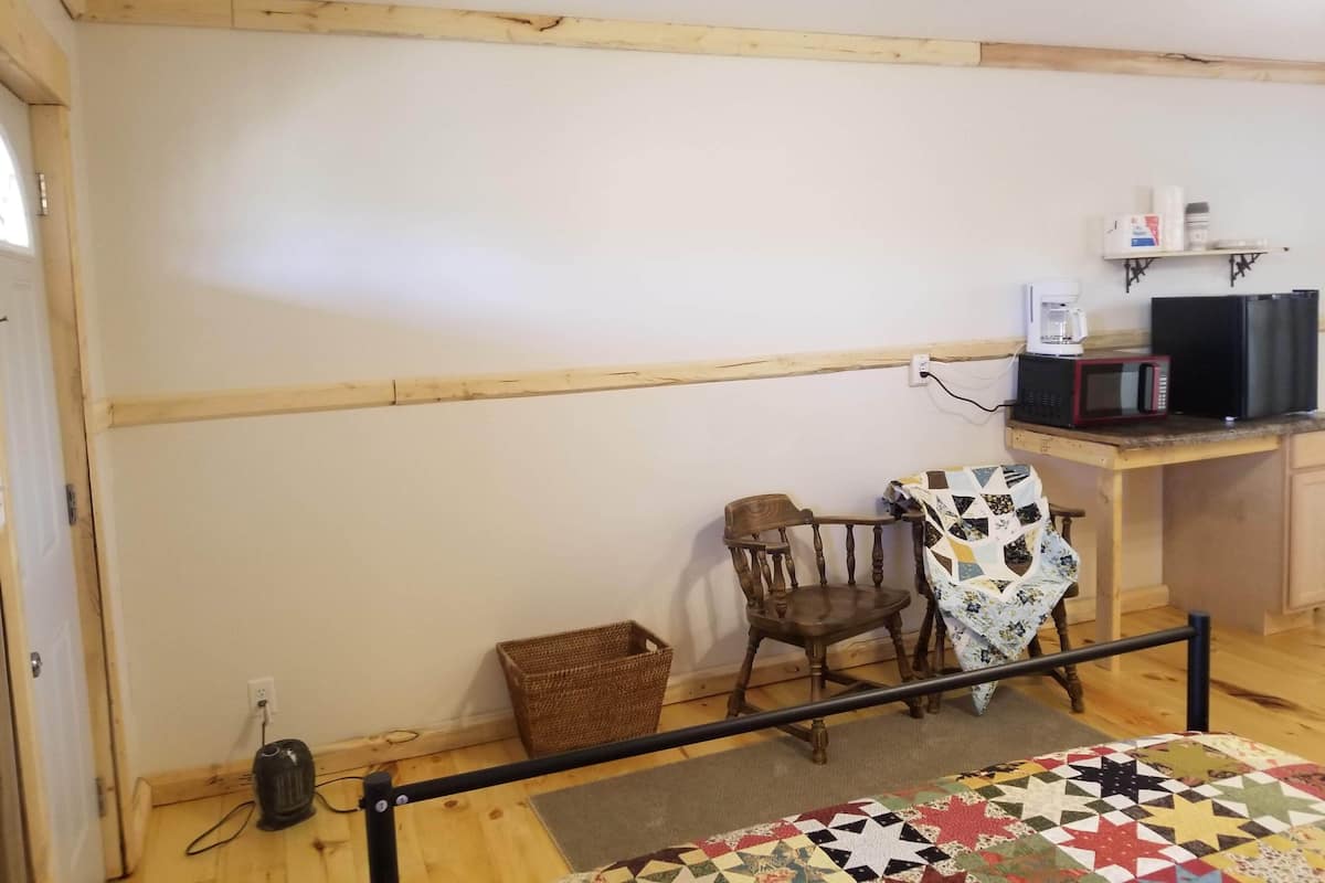 A room with a wooden floor, a desk with a computer and a microwave, a chair, a small basket, and a quilt on the floor.