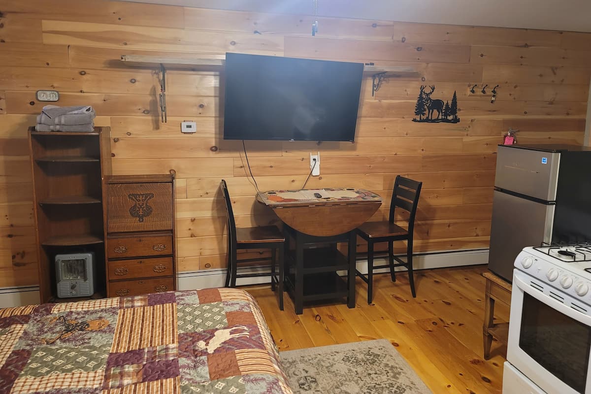 A cozy room with wooden walls, a flat-screen TV mounted above a wooden table, a refrigerator, and a bed with a patterned quilt.