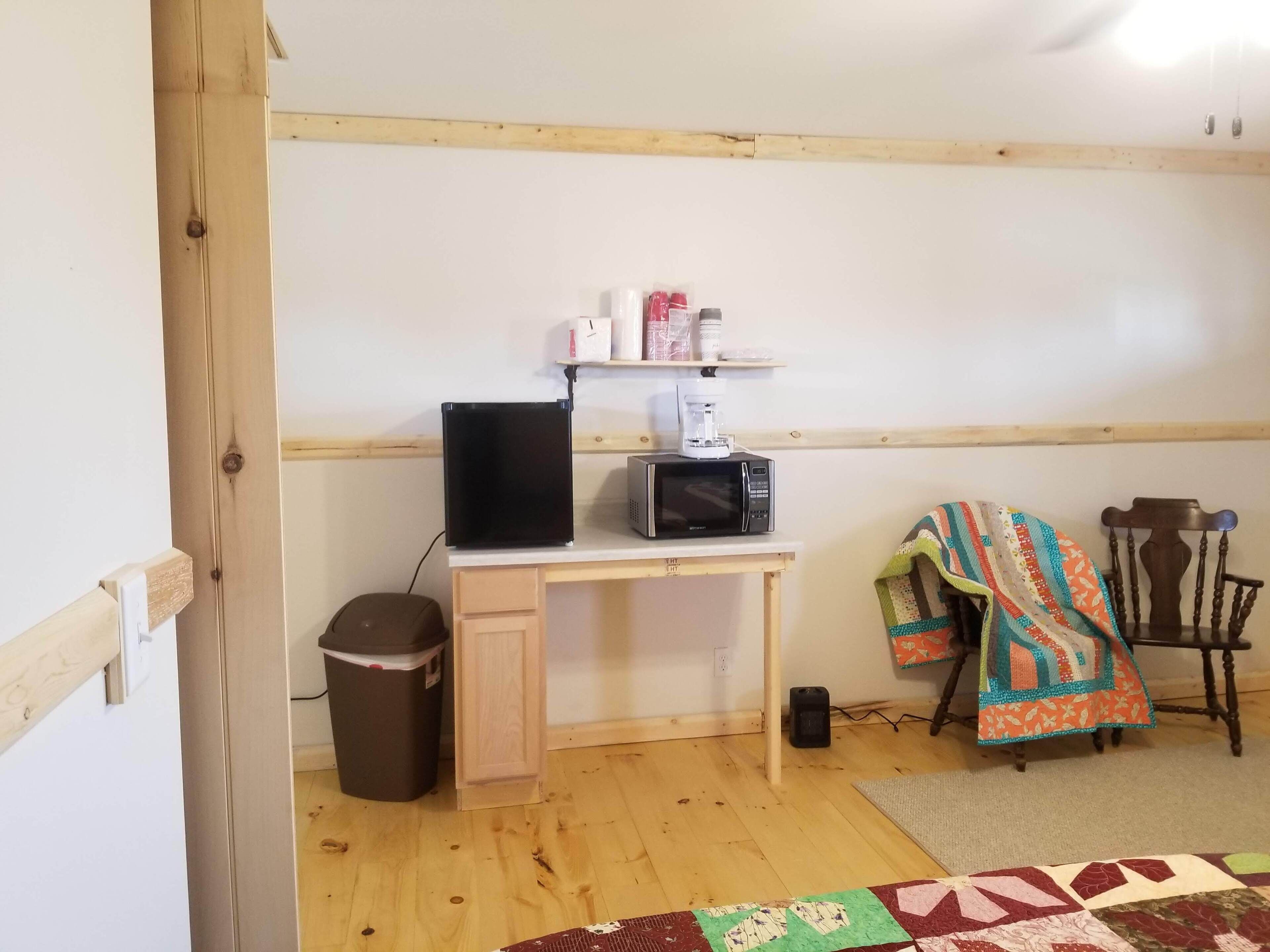 Een kleine, eenvoudig ingerichte kamer met een houten bureau, een zwarte televisie, een magnetron, een prullenbak en een stoel met een kleurrijke deken.