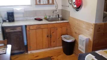 Eine kleine Küche mit Holzausstattung, einem Spülbecken und einer Arbeitsplatte. Auf dem Boden steht ein Mülleimer und ein Heizgerät.