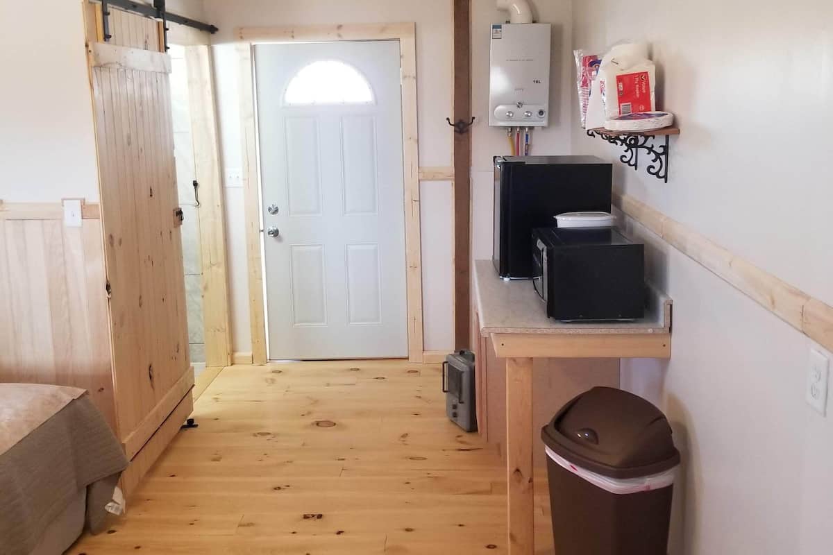 A compact room with a wooden floor, a sliding door, a ceiling fan, a small table with a computer, a trash bin, and a shelf with bags.