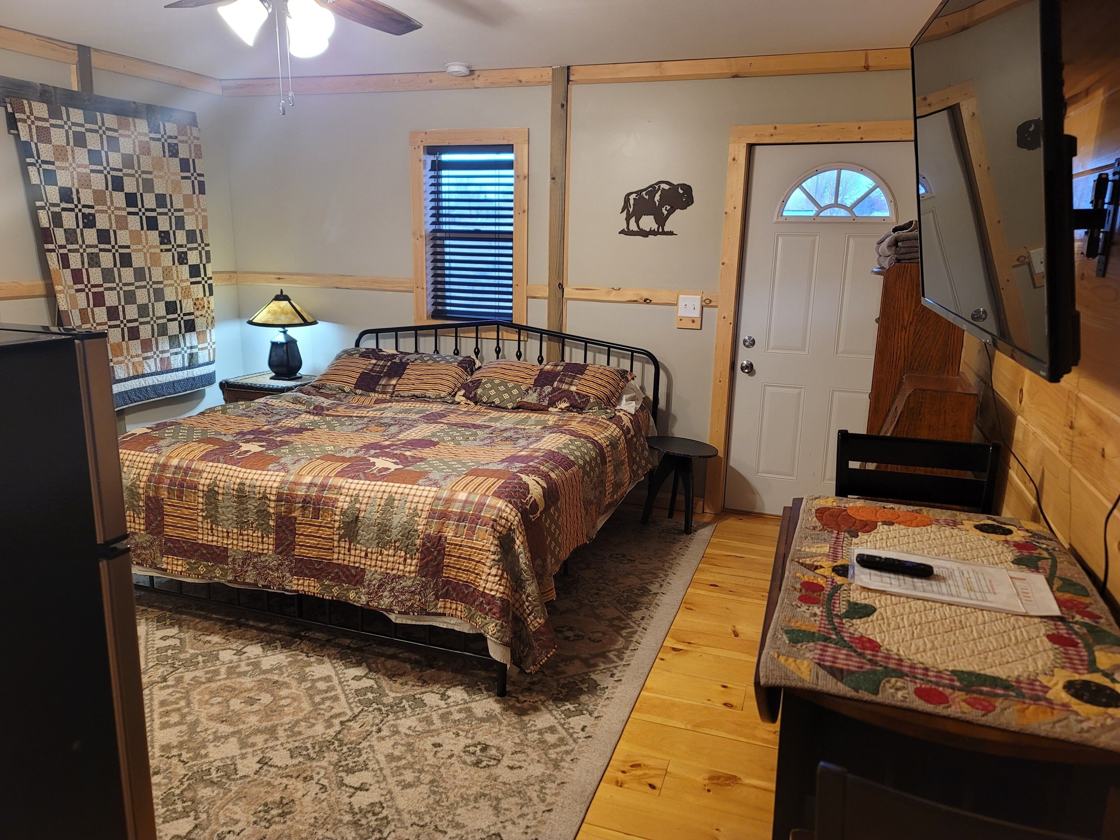 A cozy bedroom with a large bed, a desk with a chair, a lamp, a quilted wall hanging, a door with a window, and a ceiling fan.