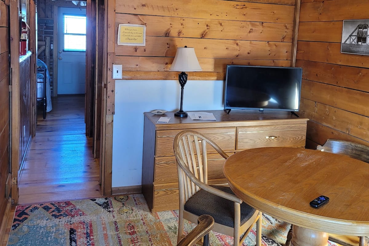 A wooden cabin interior with a dining area, a desk with a lamp, a television, and a rug.