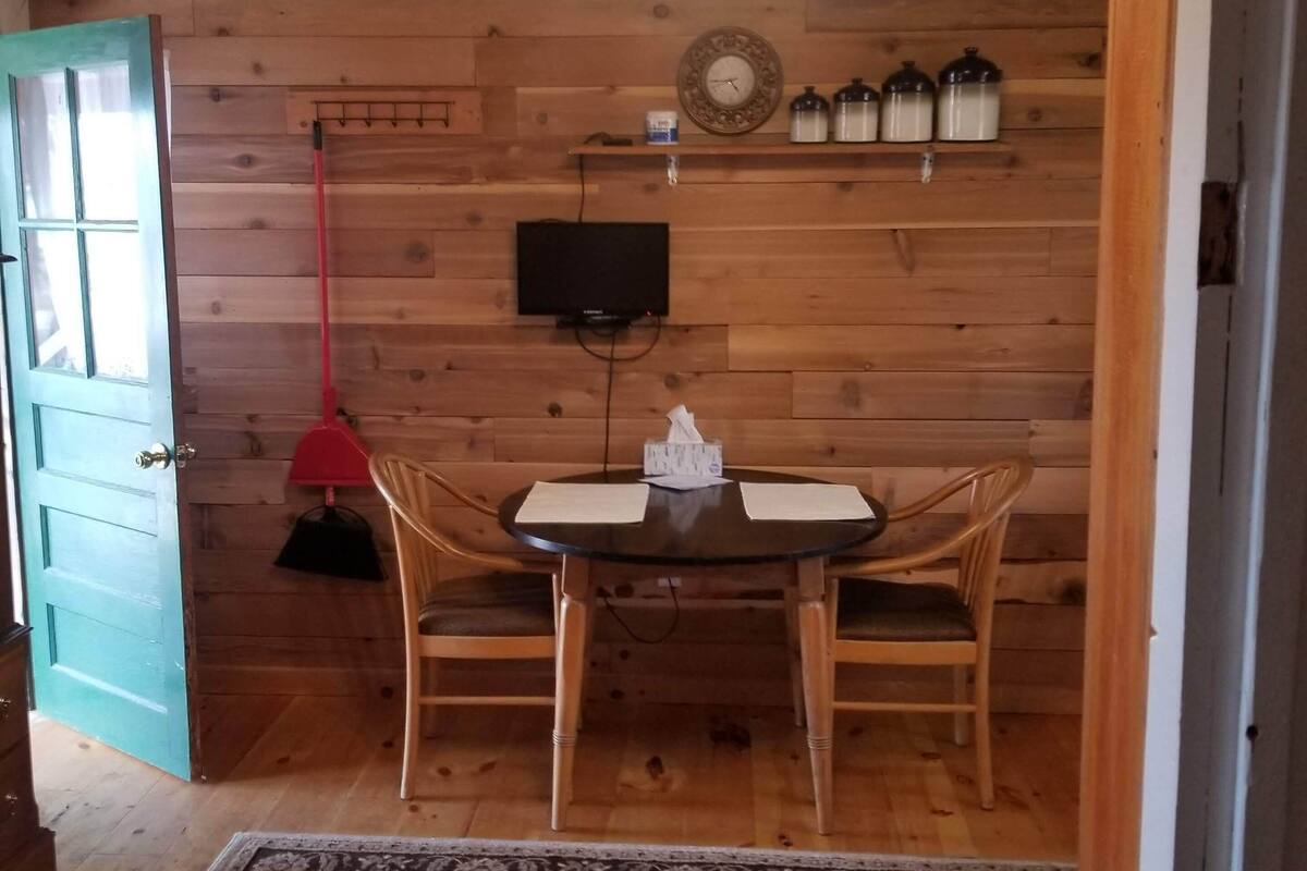 A cozy room with a wooden wall, a round table with chairs, a television, and a shelf with jars.