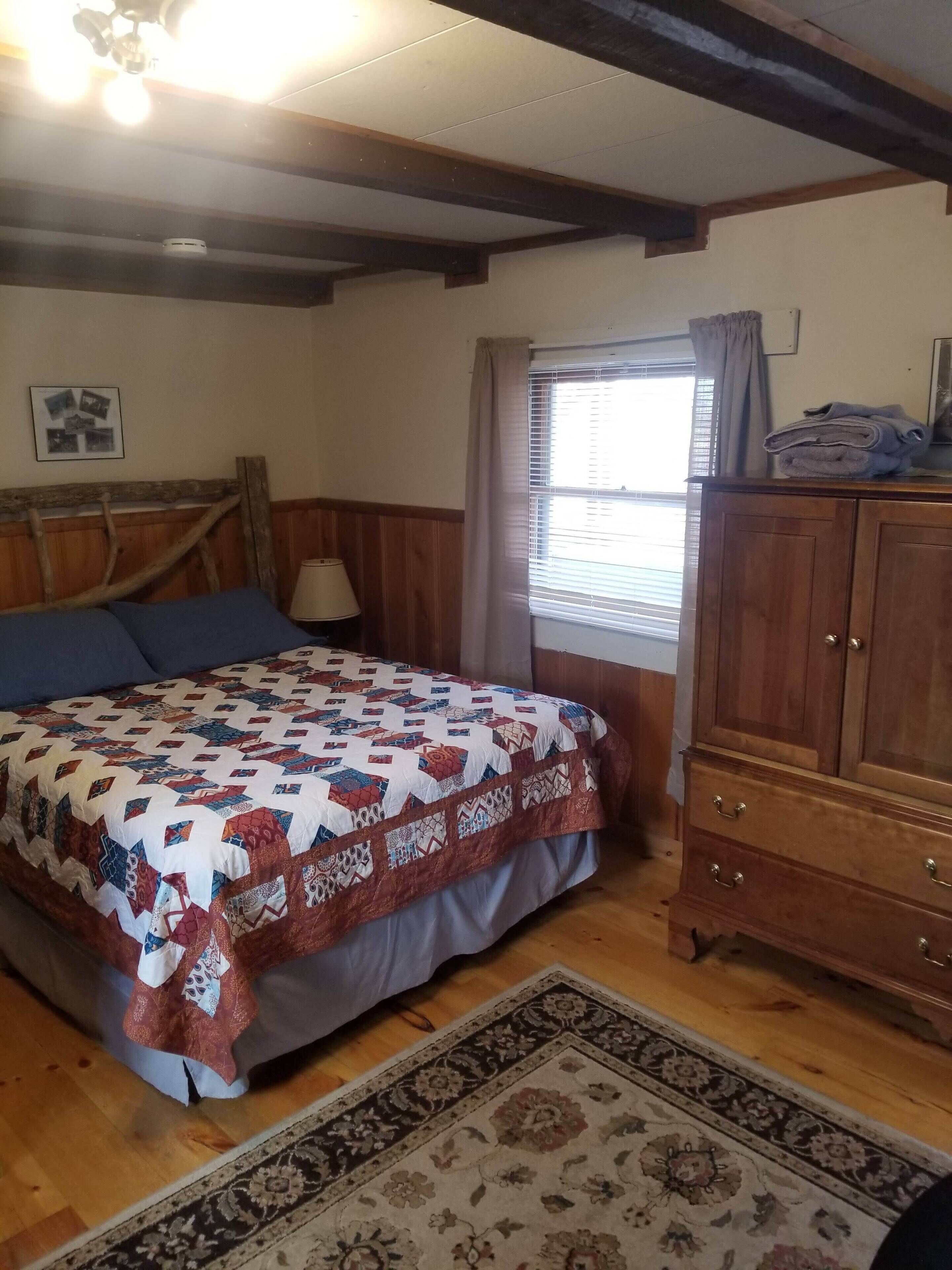 A bedroom with a bed, a wooden headboard, a nightstand, a lamp, a window with curtains, a wooden wardrobe, and a patterned rug.