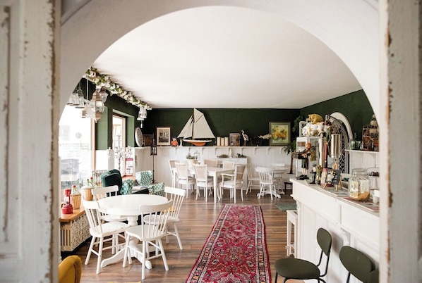 A cozy dining area with white chairs and a red patterned rug, a white archway, and a kitchen area with a green wall and various decorations.