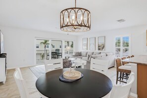 A modern living room with a round dining table, white sofas, and a large window with a view of palm trees. - Captains Cove 401~heated pool~beach access (Indian Shores)