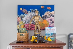 Un escritorio con una figurita de robot, un control remoto, un libro titulado 'Swan Harbor' y una cesta tejida con libros.