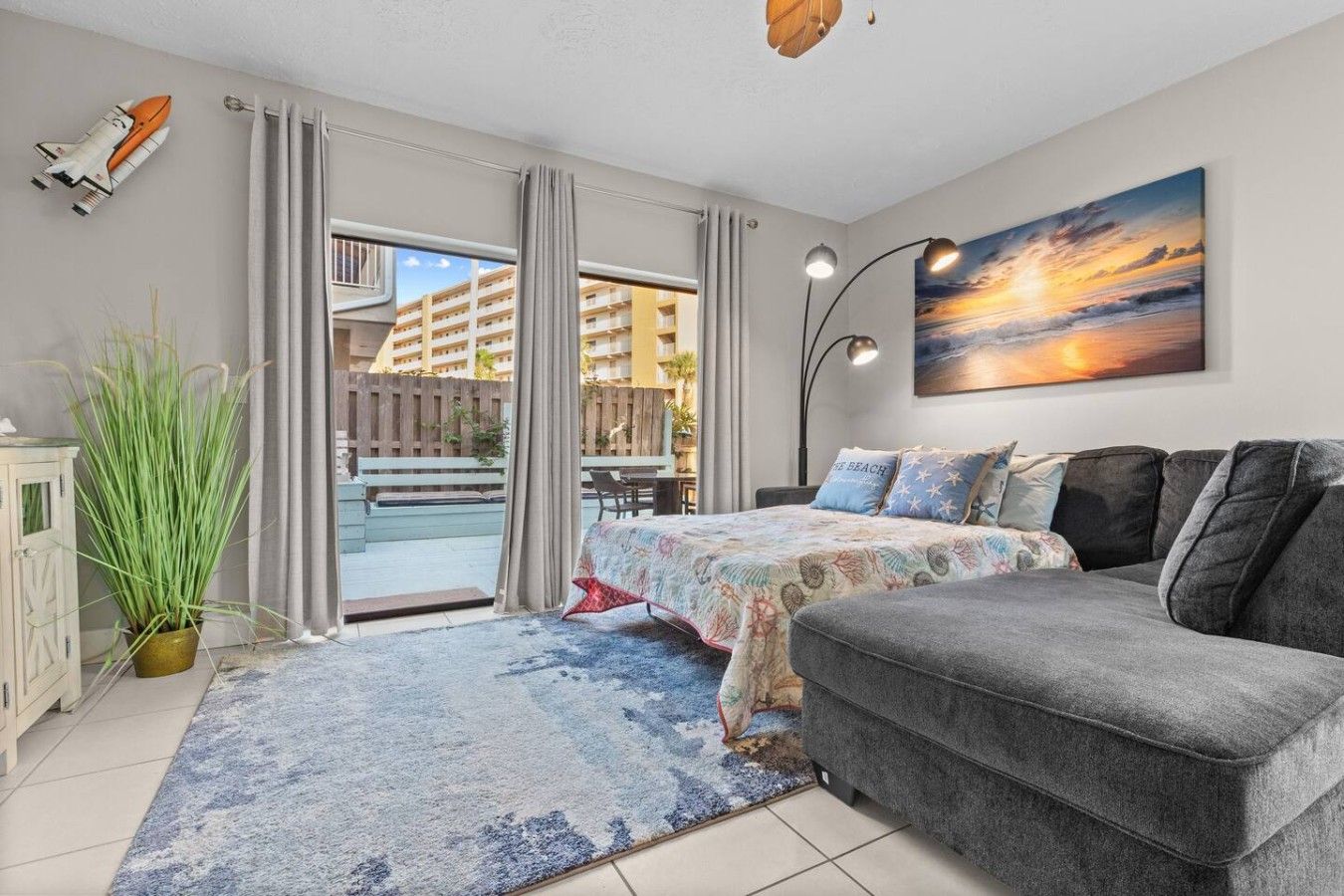 A modern bedroom with a bed, a grey sofa, a large window with curtains, a potted plant, and a painting of a sunset.