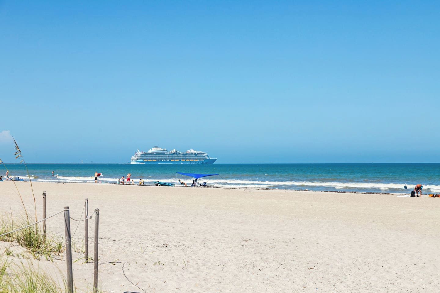 A beach with a large cruise ship in the distance, sandy shore, and people enjoying the beach.