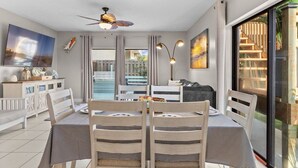 Un coin repas avec une table, des chaises et un ventilateur de plafond. On aperçoit un canapé et un téléviseur au fond.